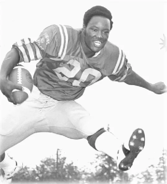 Black and white photo of a football player in uniform mid-air leaping forward while holding a football.