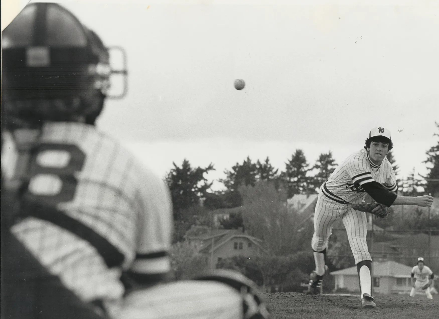 Baseball pitcher in striped uniform throwing a pitch toward a catcher wearing a helmet, with trees and houses in the background.