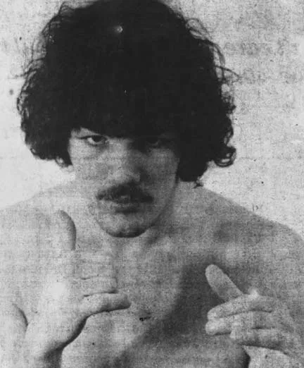 Black and white photo of a shirtless man with curly hair and a mustache making a hand gesture.
