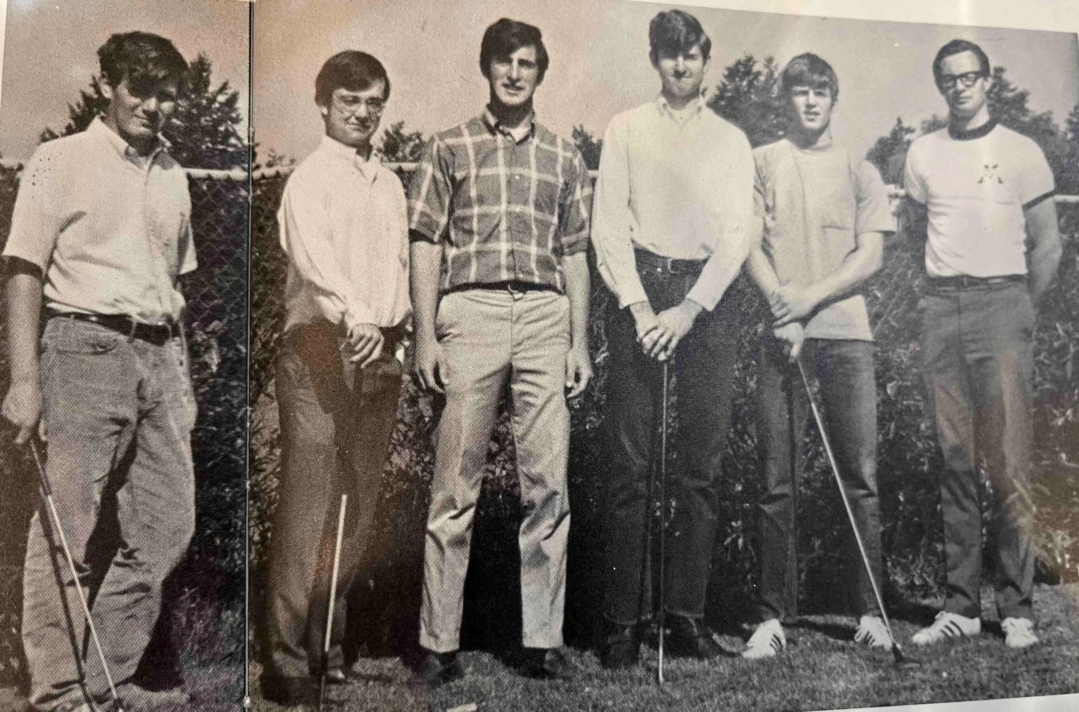 Black-and-white photo of six young men standing outdoors holding golf clubs, dressed in casual 1970s clothing.