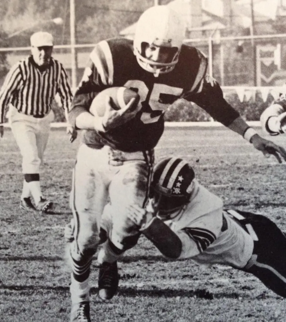 Black-and-white image of a football player wearing number 25 running with the ball while another player tackles him near the ground.