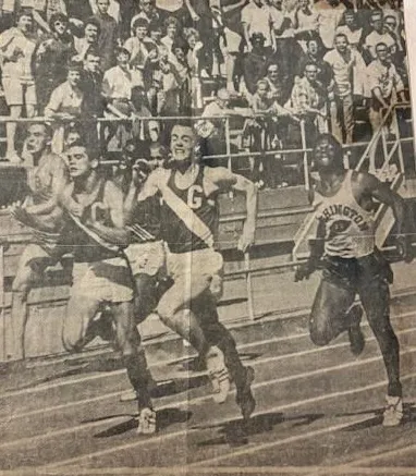 Three male athletes sprinting on a track with spectators watching from bleachers.