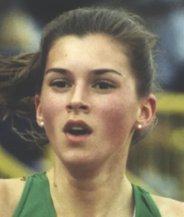 Close-up of a young female athlete running, wearing a green tank top and pearl earrings.