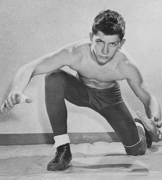 Black-and-white photo of a young male wrestler in a crouched stance, wearing wrestling shoes, tights, and shorts.