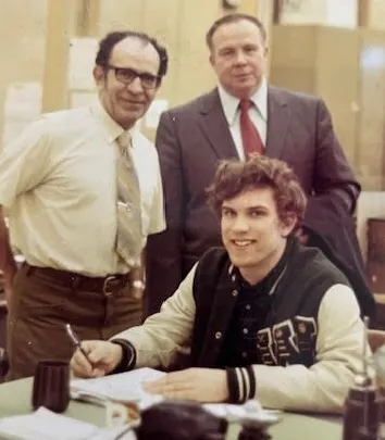 Three men in an office setting; one young man seated and smiling at a desk with papers, and two older men standing behind him, one in a suit and the other in a shirt and tie.