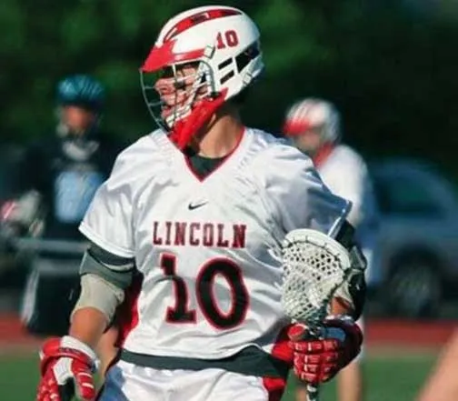 Lacrosse player wearing white Lincoln jersey with number 10 and red helmet holding a lacrosse stick.