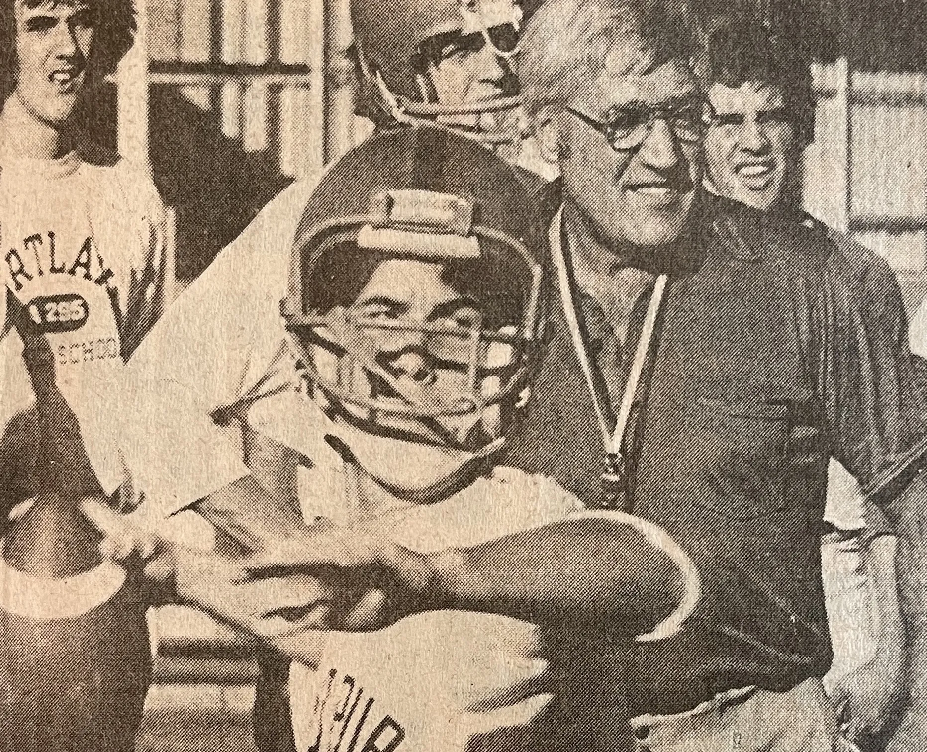 Vintage photo of a smiling football player in helmet throwing a football, with a coach wearing a whistle and others in the background.