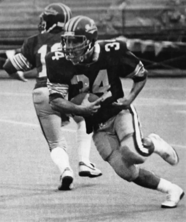 Black and white photo of an American football player wearing a number 34 jersey running with the ball on the field while another player in the background wears a number 15 jersey.