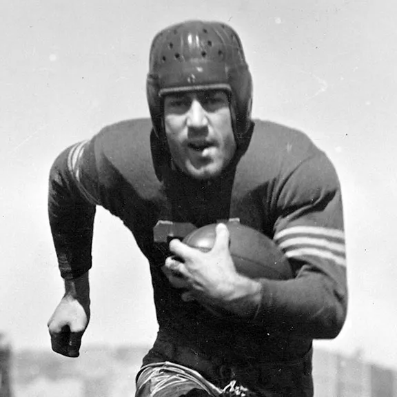 Black and white photo of a football player wearing vintage leather helmet and striped jersey running forward while holding a football.
