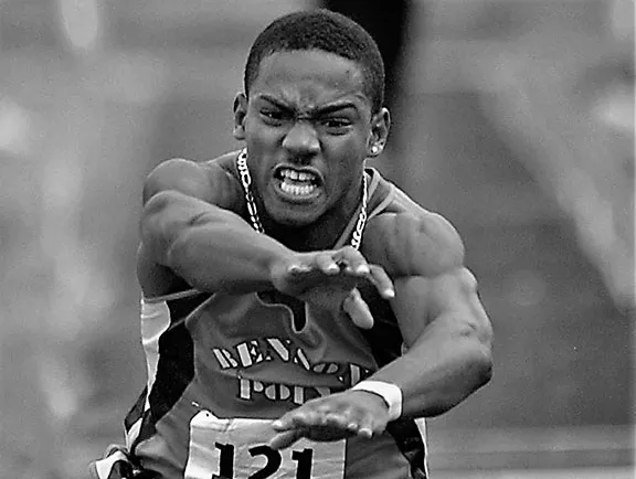 Determined male athlete mid-jump in a track and field event wearing a numbered jersey.