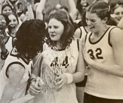 Three female basketball players celebrating; one wears a net around her neck as a trophy.