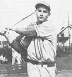 Black and white photo of a man in vintage baseball uniform swinging a bat on a field with a house in the background.