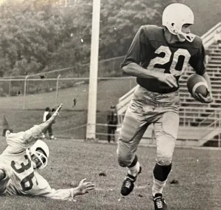 Vintage black and white photo of a football player wearing jersey number 20 running with the ball while player number 36 falls on the grass behind him.