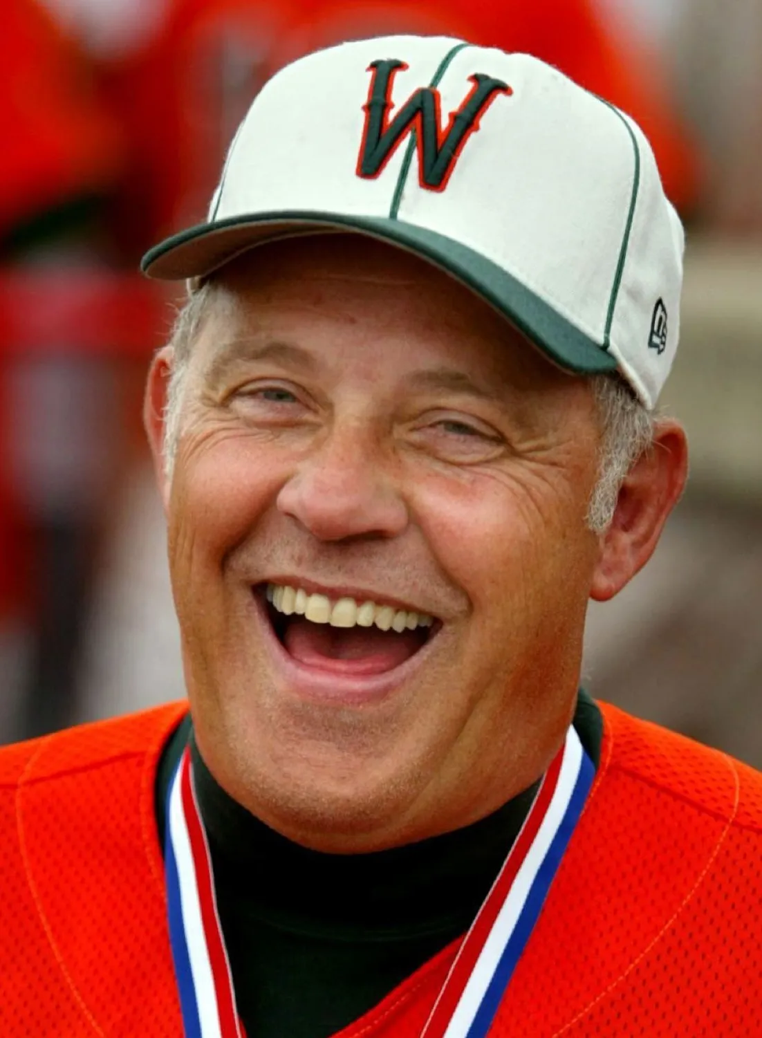 Smiling older man wearing a white baseball cap with a red and black 'W' and an orange jersey with a medal around his neck.