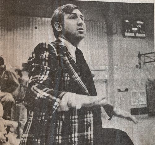 Man in a plaid jacket speaking or gesturing with his hands in an indoor setting with people in the background.