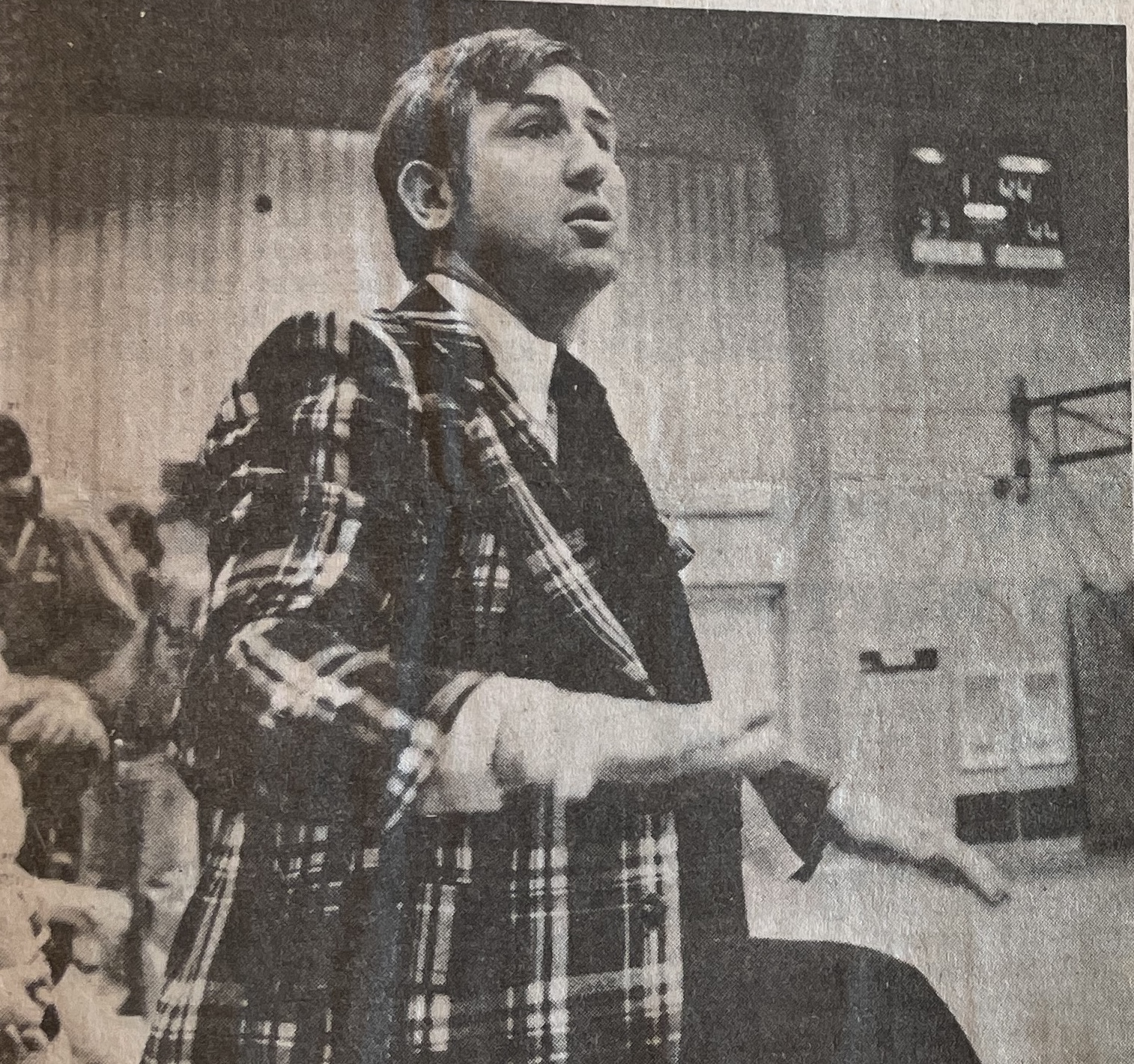 Man in a plaid jacket speaking or gesturing with his hands in an indoor setting with people in the background.