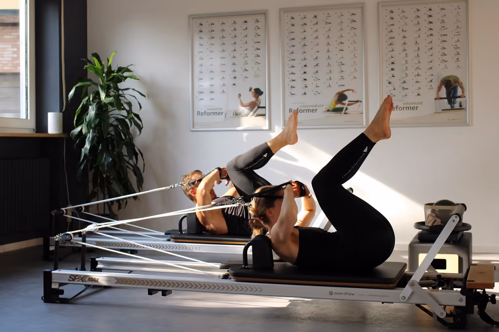Zwei Personen trainieren an Pilates-Reformer-Geräten in einem hellen Raum mit Anleitungsplakaten und einer großen Pflanze.
