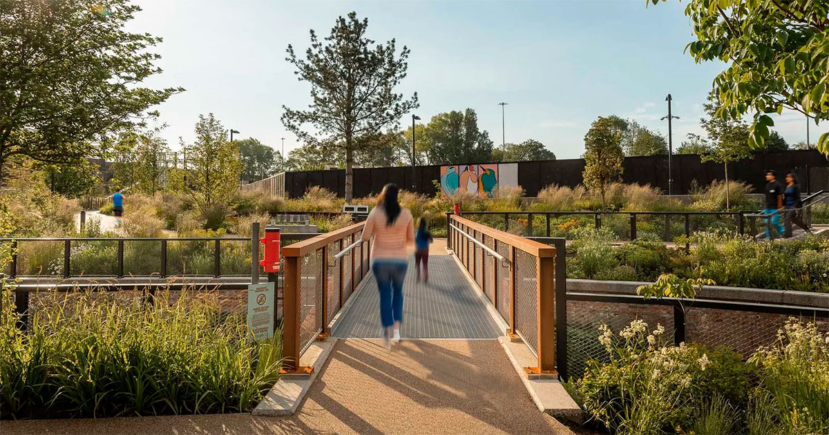 Woman pushing a pram while holding a child’s hand walking along a paved path in a sunny park with trees and grass.