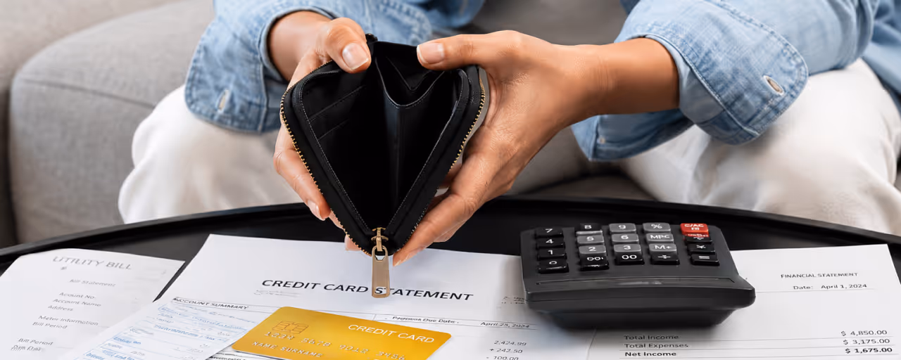Person holding an empty black wallet over credit card, utility bill, and calculator on a table.