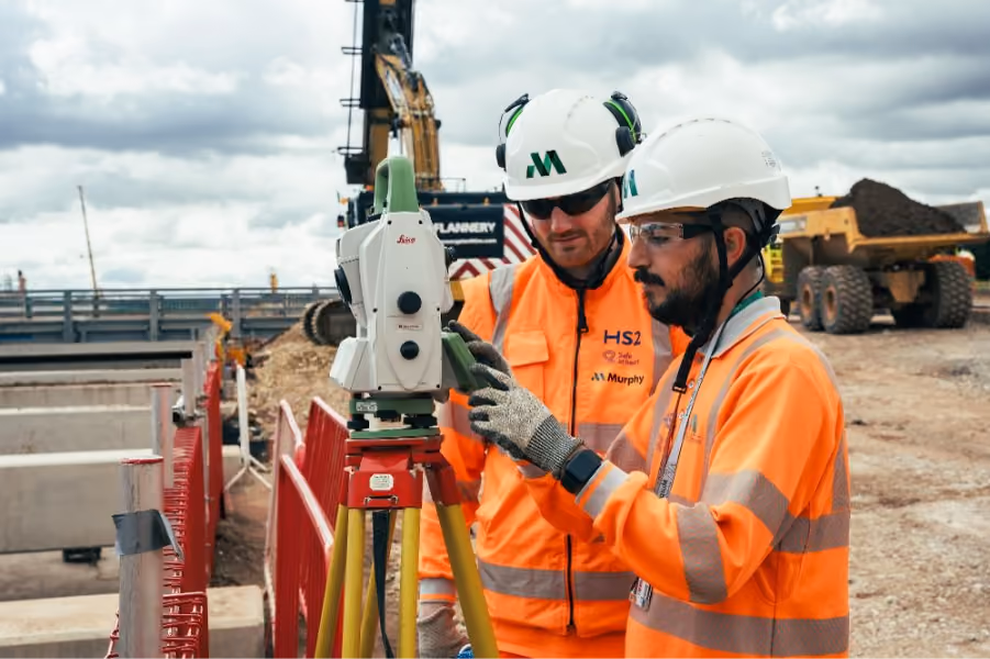 Two construction workers in safety gear operating surveying equipment on a worksite with heavy machinery.