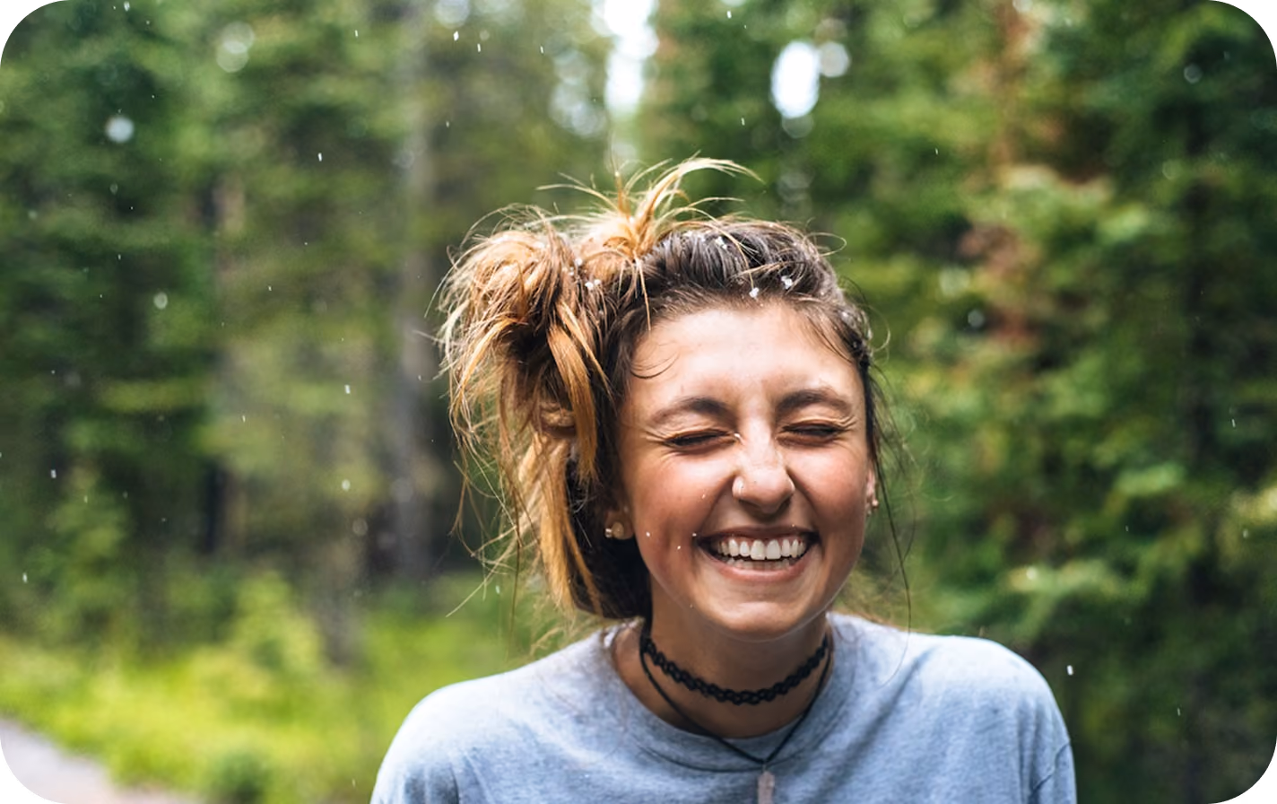 Young woman outside with eyes closed and big smile, hair in messy bun, wearing a gray shirt and choker necklace, with green trees in the background and light rain or snow falling.