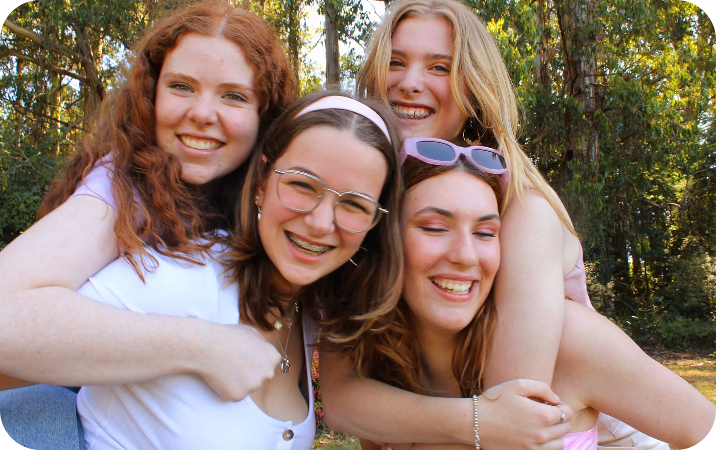 Four teenage girls outdoors smiling and hugging each other warmly in a wooded area with sunlight filtering through trees.