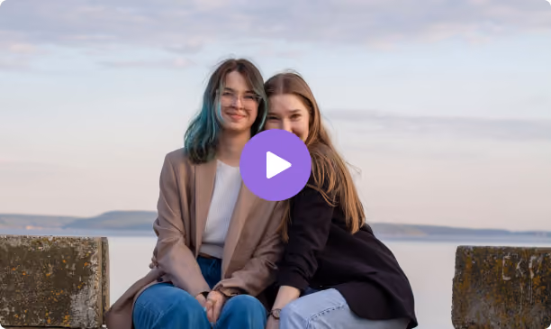 Two young women sitting close together outdoors by a body of water, smiling at the camera under a partly cloudy sky.
