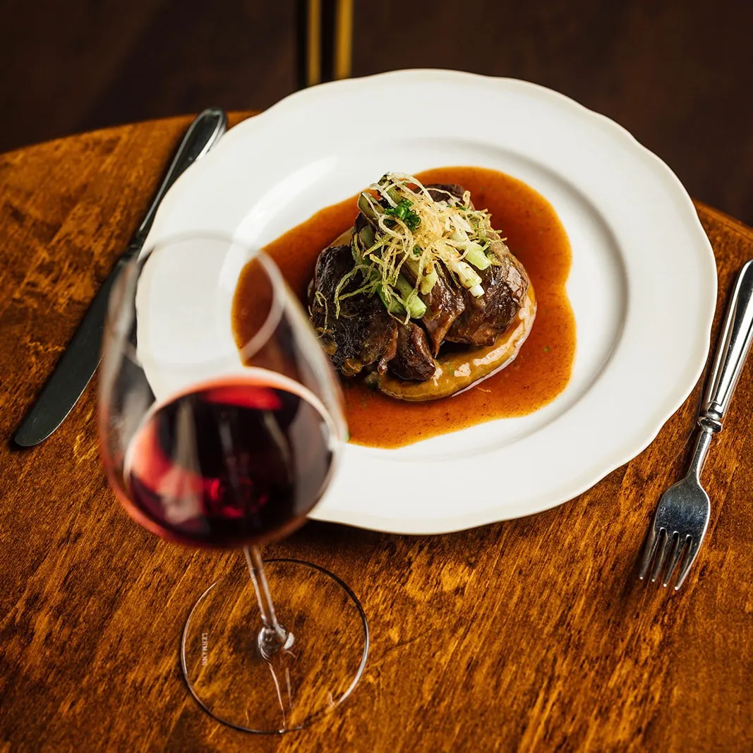 A plated gourmet beef steak topped with green garnish and fried onion strips, served on a white plate with brown sauce, accompanied by a glass of red wine on a wooden table.