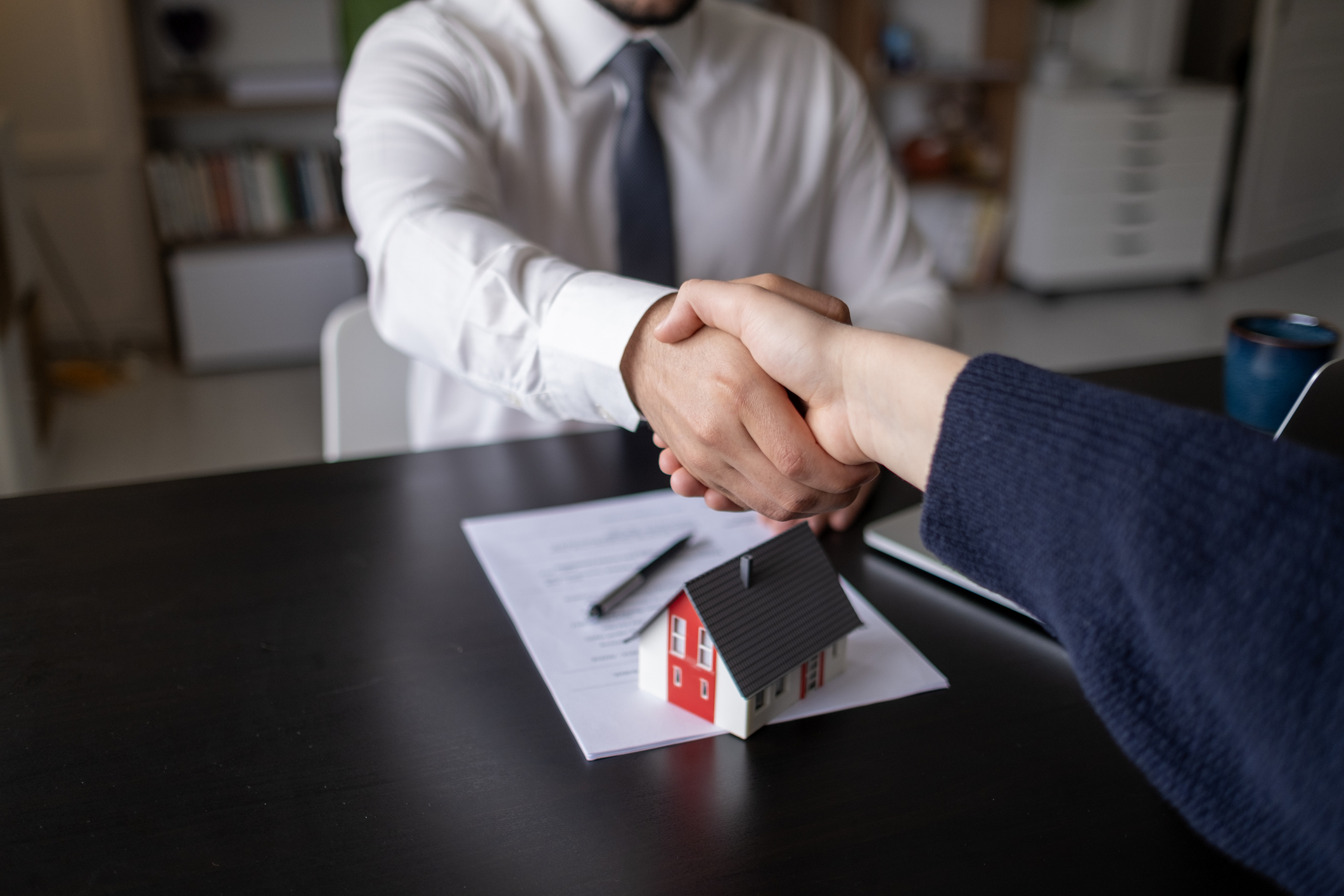 Handshake over a desk with a home model and paperwork, representing a business partnership in real estate and mortgage lending