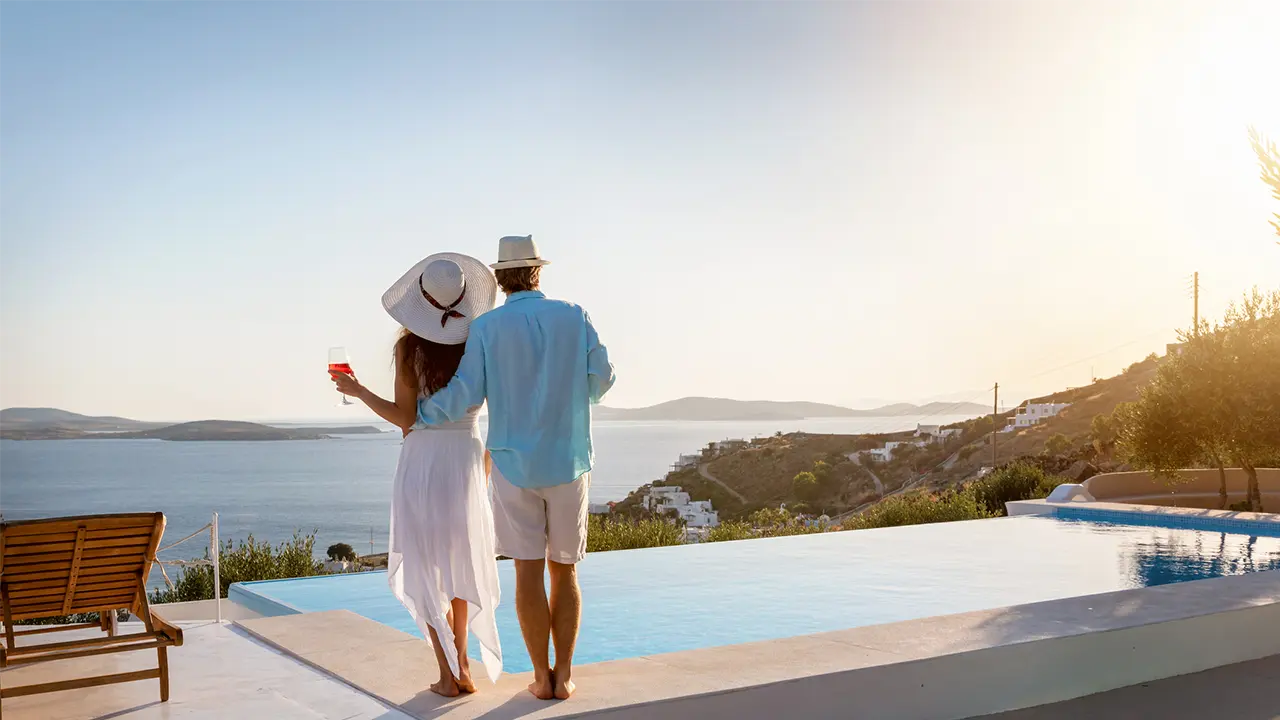 A couple standing at an infinity pool overlooking the Mediterranean at sunset, representing the identity theft risks high net worth individuals face during luxury summer travel.