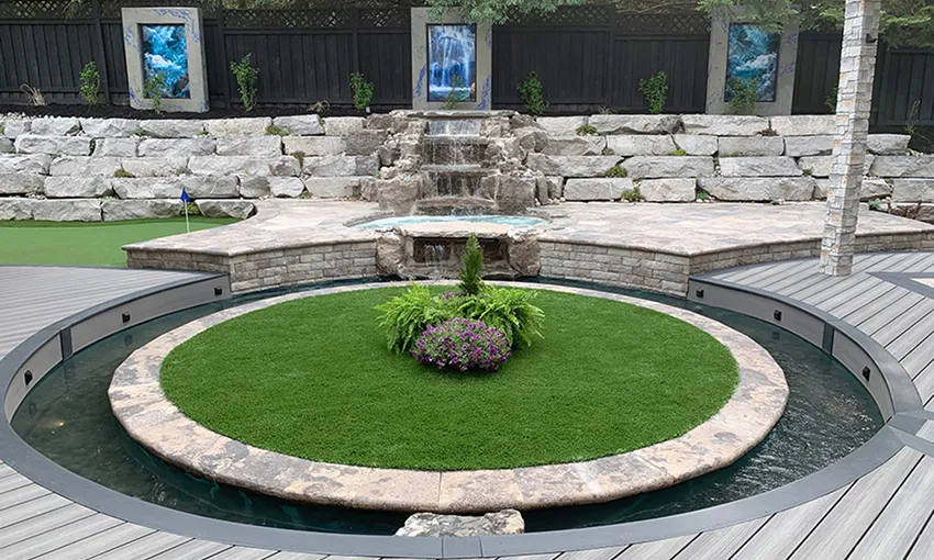 Backyard with circular stone water feature surrounding a grass and flower island, stone waterfall, and putting green behind.
