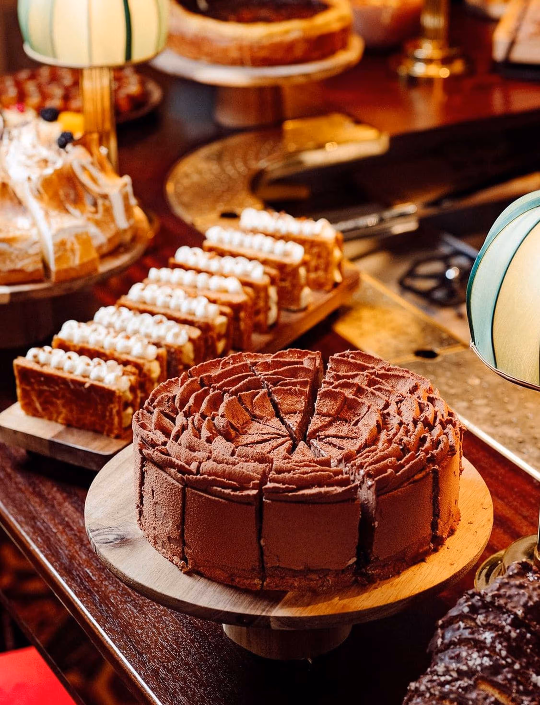 Gâteau au chocolat tranché sur un plat en bois, avec des pâtisseries garnies de crème en arrière-plan sur une table en bois.