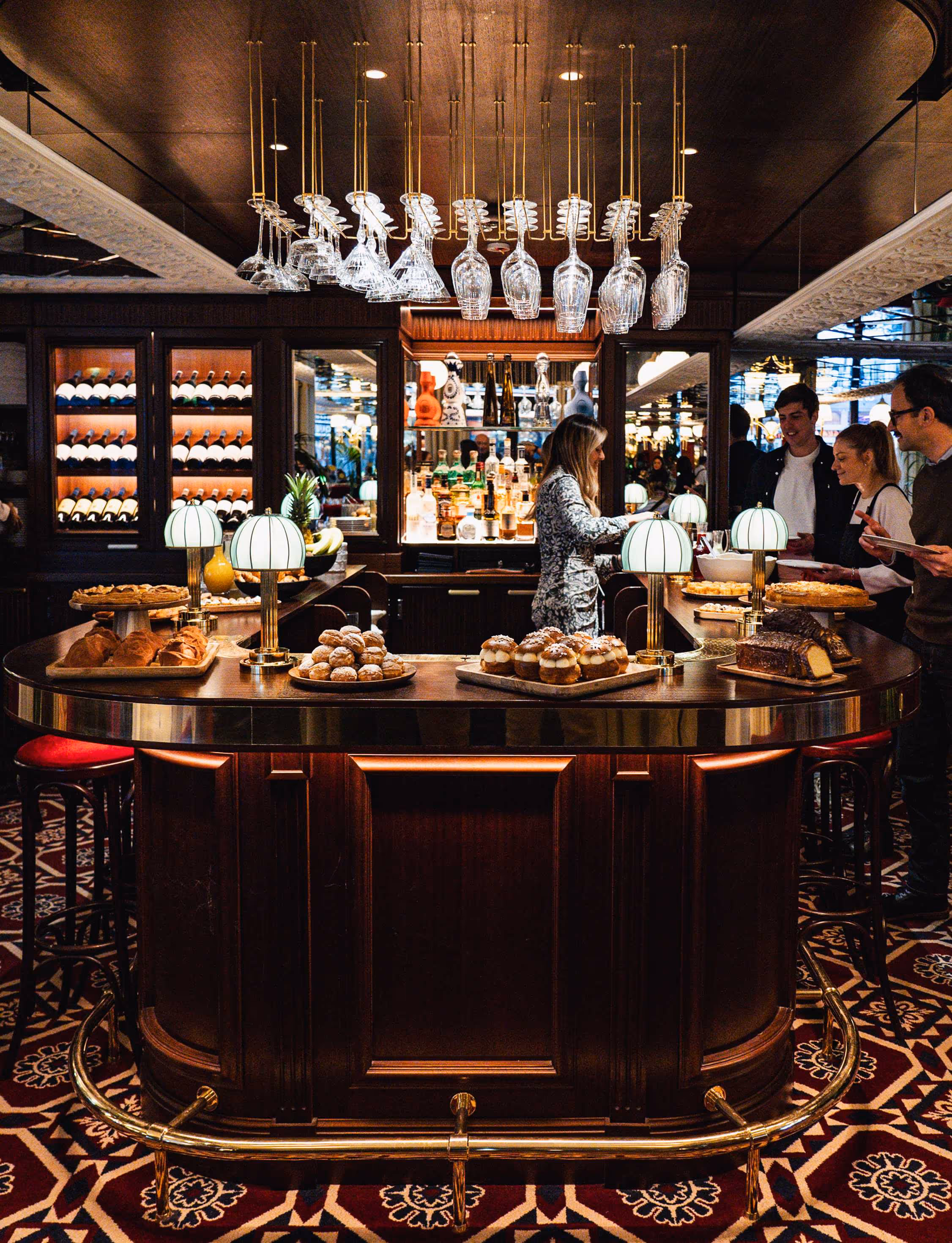 Comptoir de bar en bois avec desserts variés, verres suspendus et trois personnes prenant des pâtisseries dans un bar élégant.