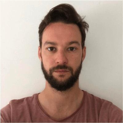 Portrait of a young man with short dark hair and a beard wearing a mauve t-shirt against a plain white background.