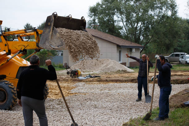 Construction crew compacting soil and adding gravel under a raised stone patio foundation