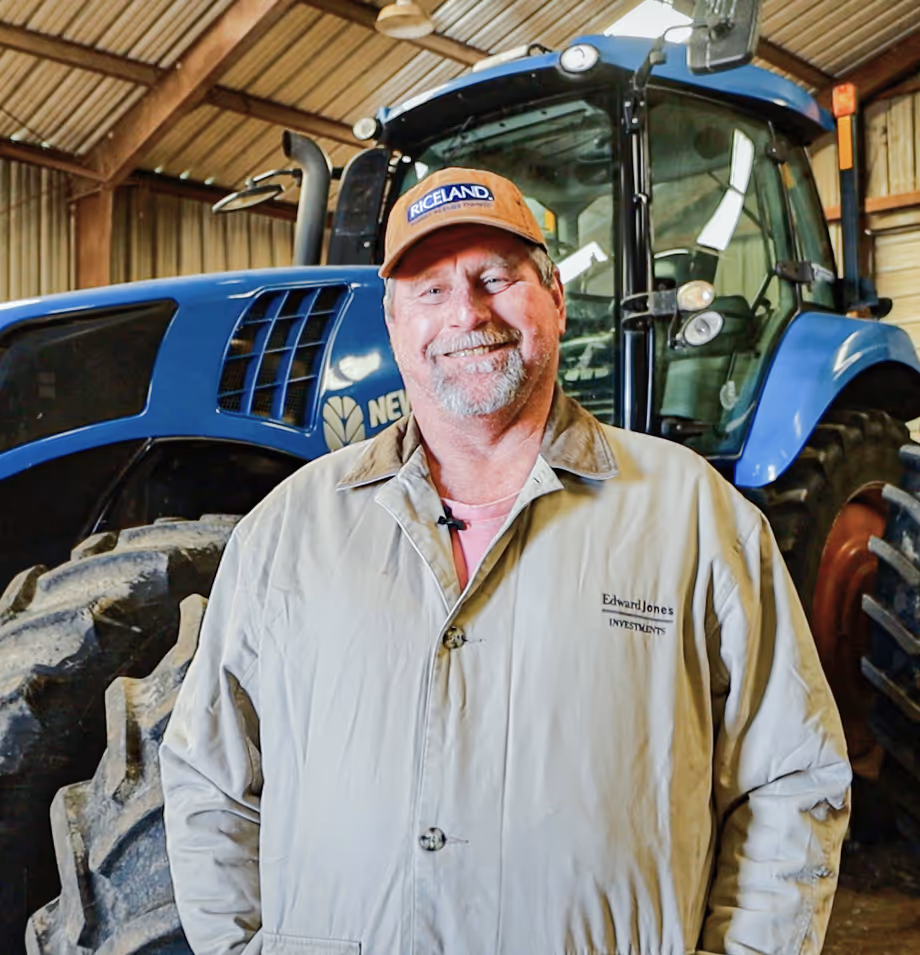 Smiling farmer in a Riceland cap standing in front of a blue tractor inside a barn.
