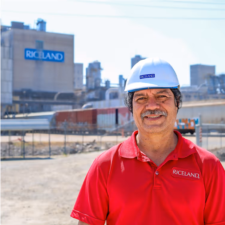 A smiling Riceland employee in a red shirt and white hard hat standing in front of Riceland grain facility.