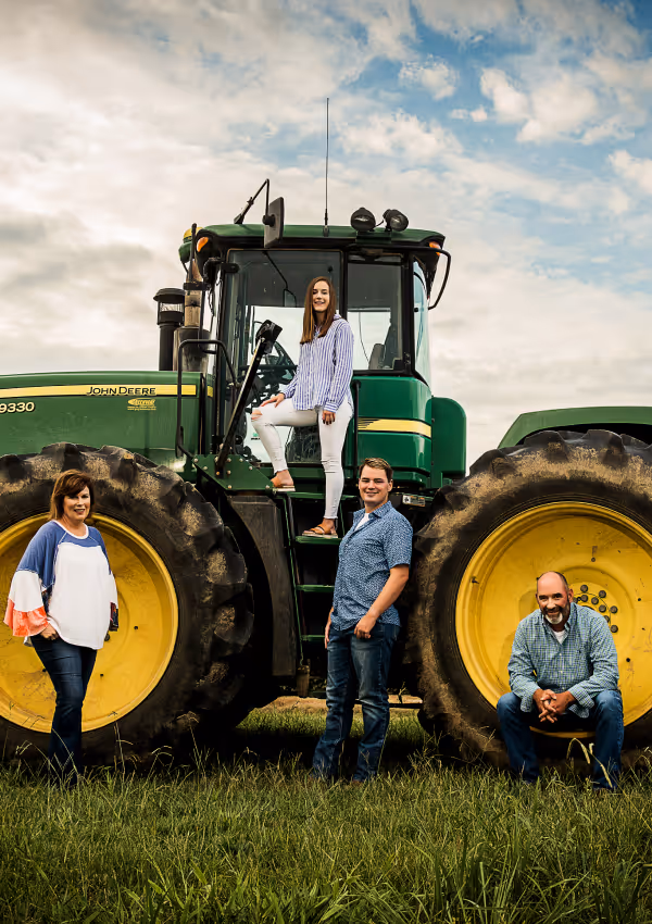 Four people posing in front of a large green John Deere tractor in a field.