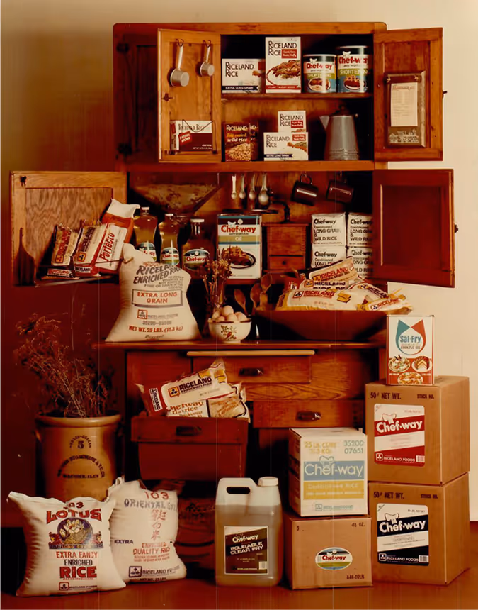 Display of Riceland and Chefway branded rice, oil, and packaged products in a wooden kitchen cabinet.