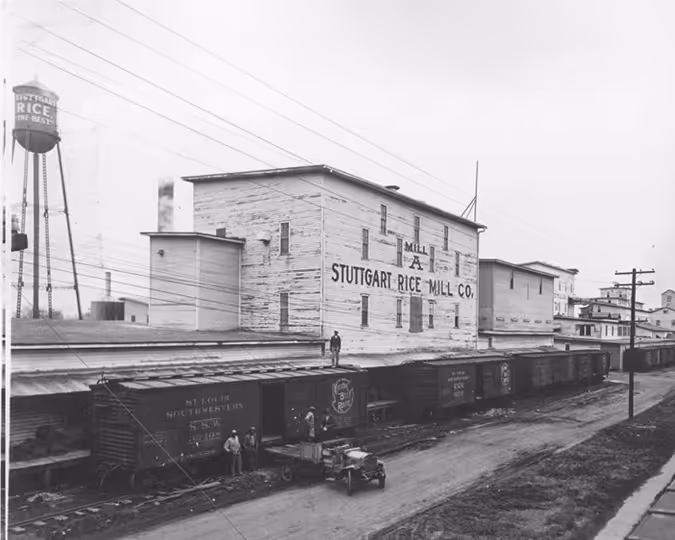 Exterior view of Stuttgart Rice Mill Company’s Mill A building, showing railroad cars and water tower.