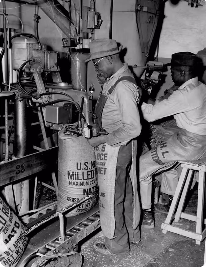 Two workers operating rice packaging machinery in a mill, wearing overalls and hats.