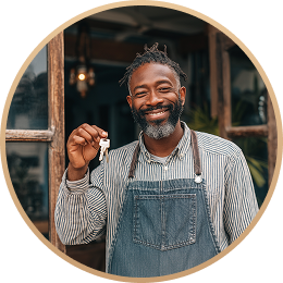 Smiling man with beard and apron holding a key in front of a doorway.