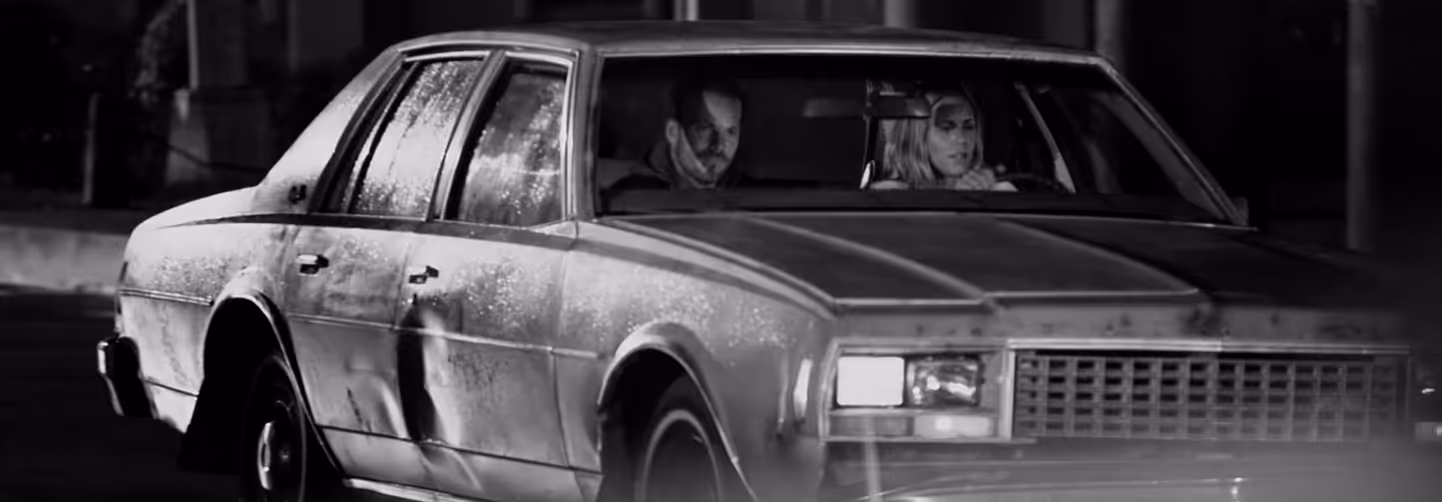Black-and-white photo of a dented, old car with two occupants inside, a man driving and a woman in the passenger seat, both looking serious.