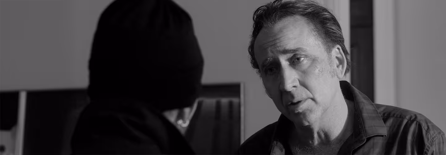 Black-and-white photo showing a serious conversation between an older man with slicked-back hair and a younger man wearing a beanie, indoors.