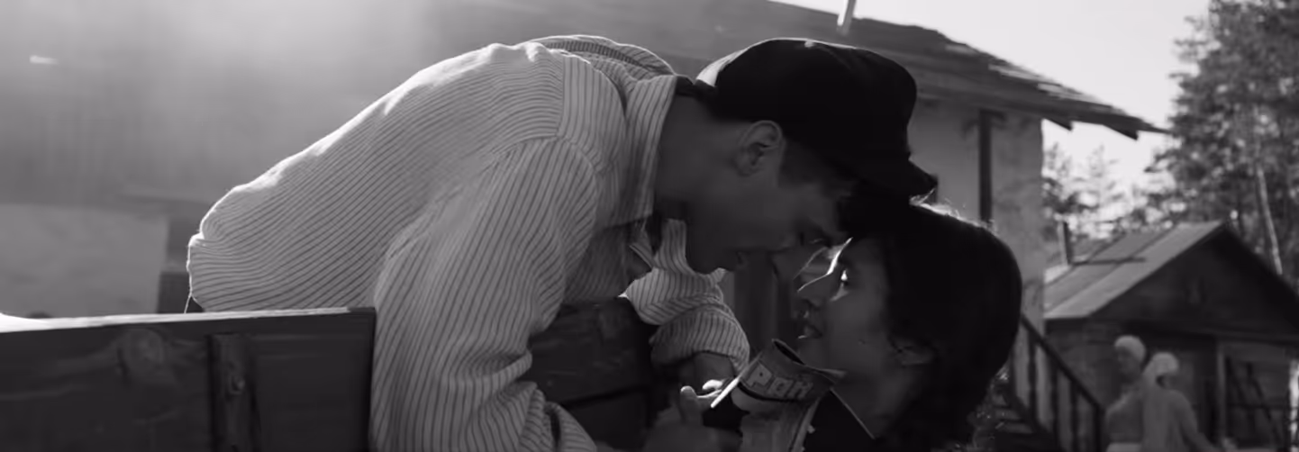 Black-and-white photo of a man and woman leaning over a wooden railing, looking into each other's eyes in a tender moment, set in a rustic outdoor environment.