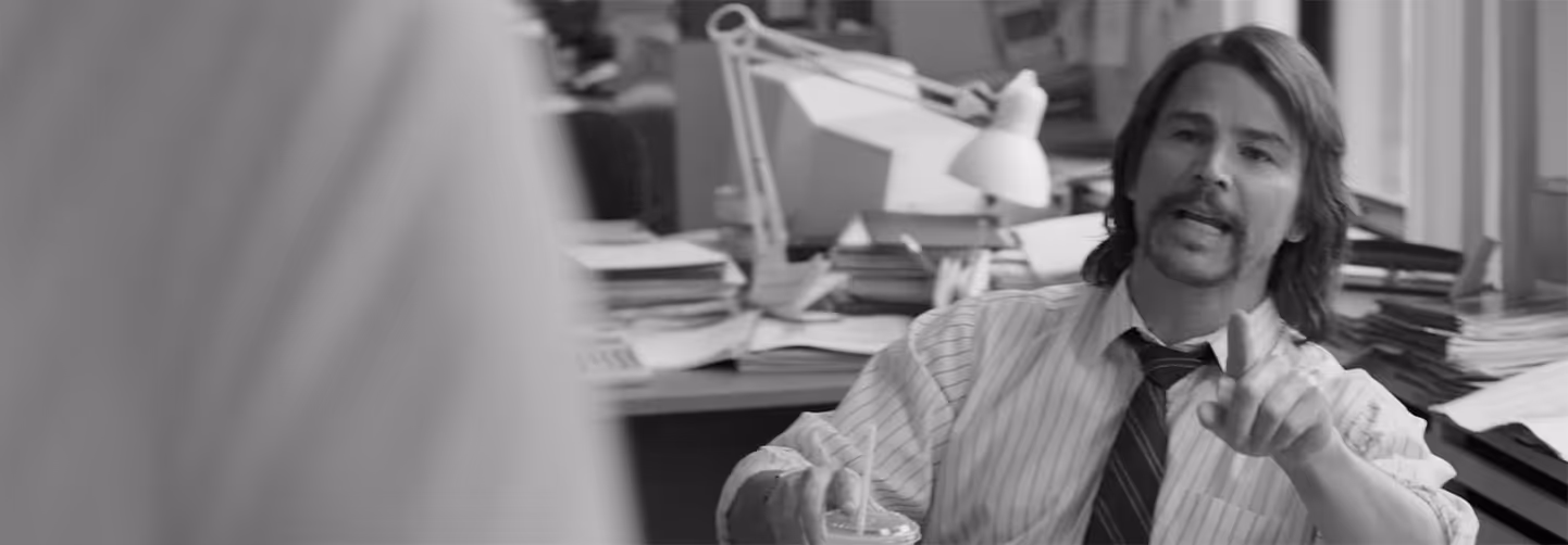 Black and white photo of a man with medium hair and mustache, sitting at a cluttered office desk, talking and gesturing.