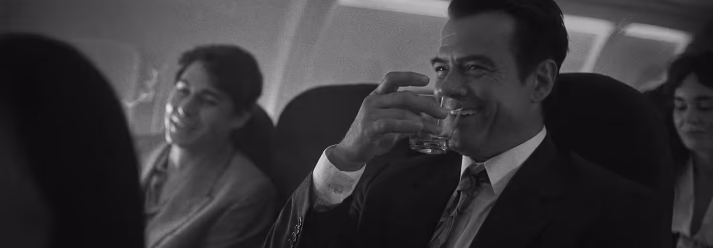 Black-and-white photo of a man in a suit smiling and holding a glass while seated inside an airplane cabin, with other passengers in the background.