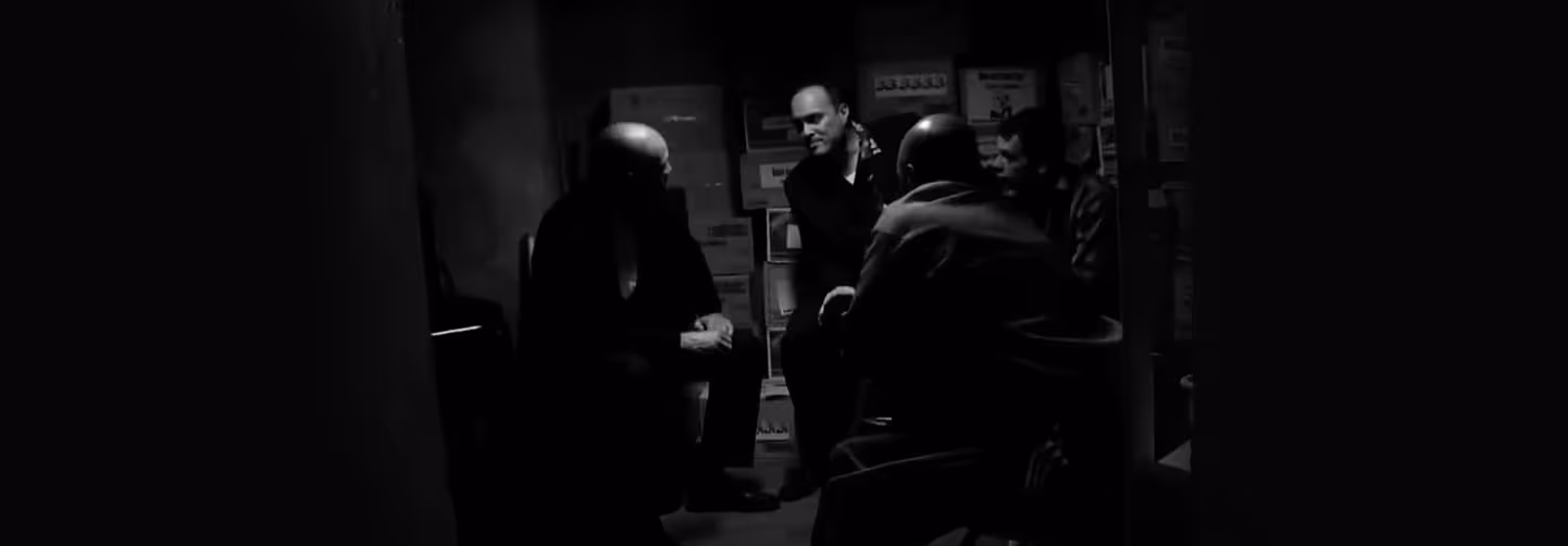 Black-and-white banner showing three people in a dimly lit storage or workroom, with one seated facing away, another standing, and the third partially visible, creating a tense atmosphere.