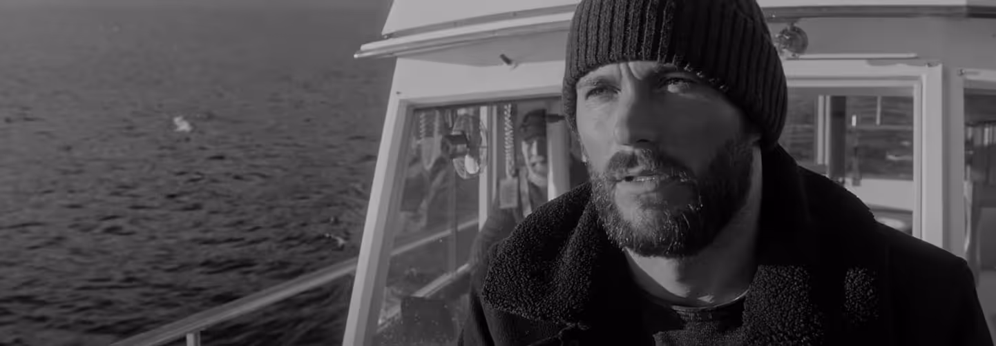 Black and white banner image of a bearded man wearing a knit hat and heavy coat, standing on a boat with water in the background, looking off to the side.