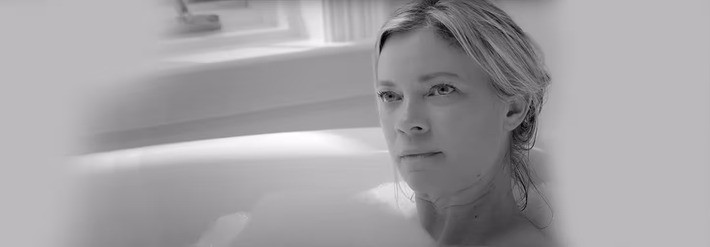 Black and white banner image of a blonde woman sitting in a bathtub, looking contemplative, illuminated by soft natural light from a window.